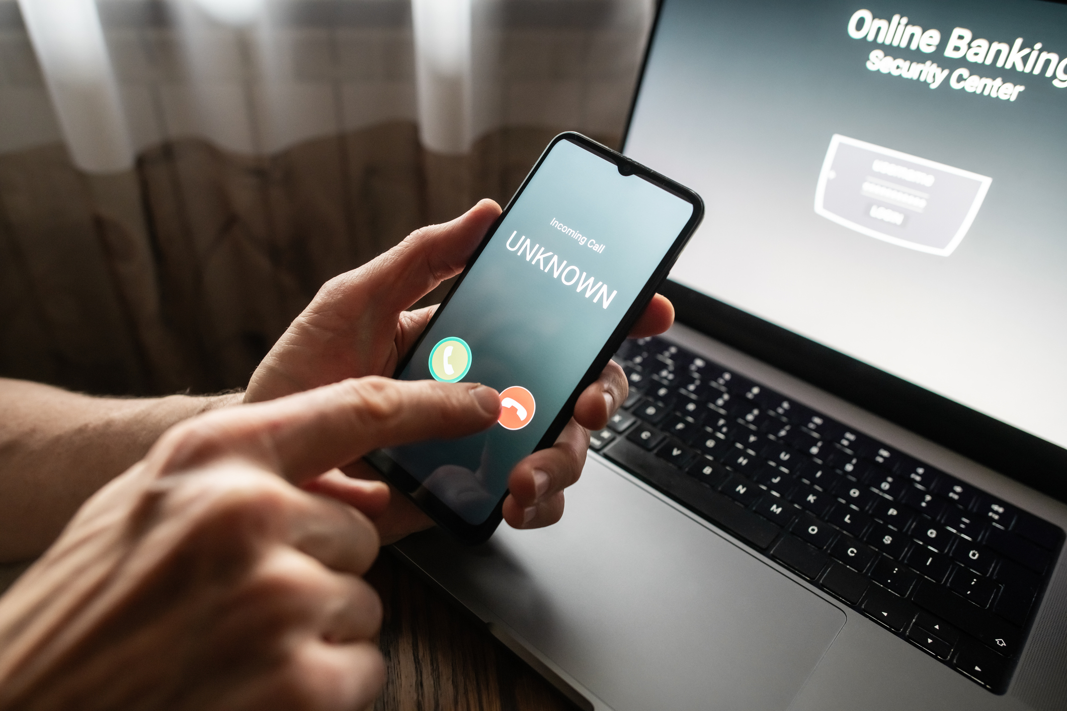person holding phone up with an unknown number calling and the online banking app on the computer screen behind the phone.